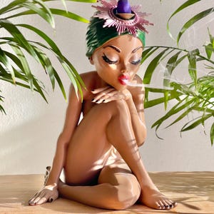 Könnte beinhalten: Nackte Figur einer Frau mit grüner Kopfbedeckung und dekorativem Kopfschmuck. Die Figur raucht, Rauch kommt aus ihrem Mund. Die Frau sitzt mit verschränkten Beinen und geschlossenen Augen. Die Figur ist von grünen Pflanzen umgeben.