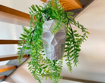 Puede incluir: Una maceta blanca con forma de calavera geométrica cuelga de una cuerda blanca, llena de vibrantes plantas de helecho artificial verde. La maceta tiene un diseño angular moderno. Las hojas de helecho caen en cascada hacia abajo.