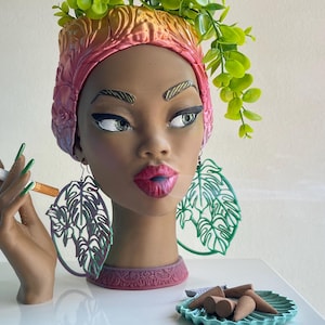 May include: A decorative head-shaped planter with a woman's face, wearing a pink and gold head wrap and large leaf earrings. The planter holds green foliage. A hand holds a lit cigarette, and smoke drifts away. A small dish holds incense cones.