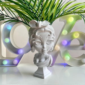 Può includere: Vaso da fiori in ceramica bianca a forma di busto di donna con un fiocco nei capelli. Il vaso è appoggiato su un tavolo davanti a un cartello luminoso colorato.