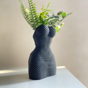 May include: A black ceramic vase shaped like a human torso with a textured surface. The vase is filled with green leafy plants.