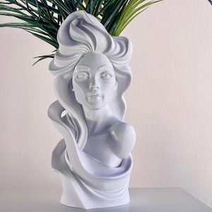 May include: A white bust vase featuring a woman's face with flowing hair and draped fabric. The vase holds green leafy stems, creating a decorative floral arrangement. The sculpture is a unique home decor item.