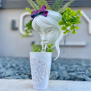 May include: White ceramic planter shaped like a woman's head with a purple bow in her hair. The planter is filled with green plants and a white cup with the text "Drink Me Coffee" is placed in front of the planter.