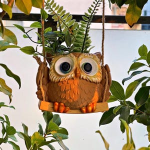 Puede incluir: Una maceta marrón con forma de búho, suspendida por una cuerda, llena de plantas verdes. El búho tiene grandes ojos azules y patas naranjas. La maceta está sobre una base de columpio de madera. El fondo está borroso con follaje verde.