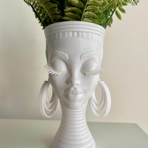 May include: White ceramic planter shaped like a woman's head with long eyelashes and large hoop earrings. The planter has a textured neck and a round base. A green leafy plant is in the planter.