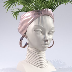 Peut inclure: Un pot en céramique blanche en forme de tête de femme avec un turban rose, des boucles d'oreilles créoles dorées et un anneau de nez doré. Le pot a un cou en couches et est rempli de feuilles de palmier vertes.