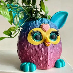 以下が含まれることがあります： 大きな目と額にピンクのハートが付いた、漫画のフクロウの形をした、紫と青のふわふわのプランター。プランターはターコイズグリーンのベースで、緑の葉の植物でいっぱいです
