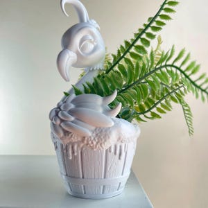 May include: A white decorative planter shaped like a cupcake with a bird on top, holding green fern-like plants. The planter has a textured, drippy frosting design and a wooden barrel-like base.