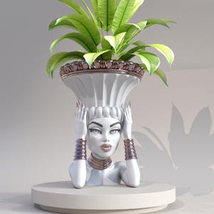 Può includere: Vaso in ceramica bianca a forma di testa di donna con una pianta verde che cresce dalla cima. Il vaso ha un bordo dorato e la donna indossa braccialetti e una collana dorati.