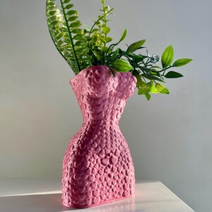 May include: A pink ceramic vase shaped like a human torso with a textured surface. The vase is filled with green leafy plants.