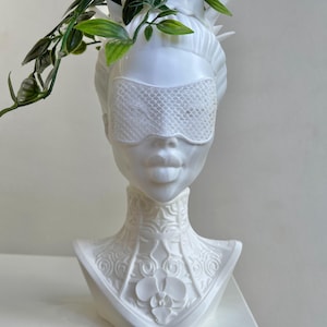 Puede incluir: Maceta de cerámica blanca con forma de busto de mujer con una máscara de encaje para los ojos y un diseño floral detallado alrededor del cuello. La maceta tiene una pequeña abertura en la parte superior para plantar.