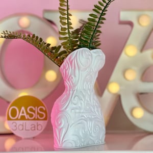 May include: White ceramic vase shaped like a woman's torso with an intricate floral design. The vase is filled with green ferns and sits in front of a white "LOVE" sign with light bulbs.
