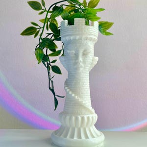 May include: White ceramic planter shaped like a castle tower with a leafy green plant growing out of the top. The planter has a detailed design and a textured surface.