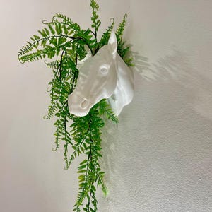 Op de afbeelding: Witte keramische paardenkop plantenbak met groene, bladerige planten die uit de bovenkant groeien.