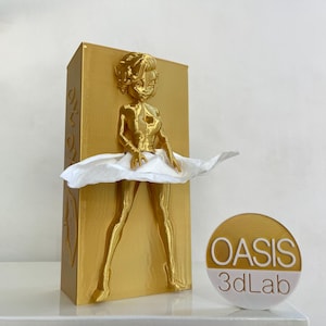 May include: Gold-colored tissue box holder with a 3D printed figure of a woman wearing a white skirt. The skirt is made of tissue paper and the figure is holding the tissue box. The tissue box holder is labeled "Oasis 3dLab".