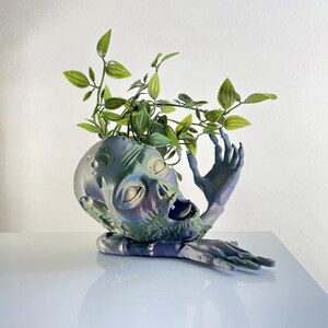 May include: A zombie-themed planter with a distressed, iridescent finish. The open-mouthed head is supported by a reaching hand and arm. Green leafy plants sprout from the top of the head, creating a unique and spooky decorative piece.