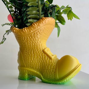 Op de afbeelding: Een gele laarsvormige plantenbak met een groene ombre-basis, gevuld met groene bladplanten.