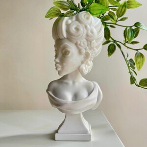 May include: A white sculpture of a woman's bust with detailed curly hair and a draped garment. The bust is on a white base, with green foliage placed in the head. The sculpture is a classic design.