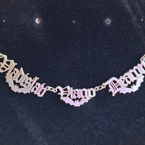 May include: A gold necklace with three nameplates in a gothic font. The nameplates read "Vladish", "Diego", and "Deacon".