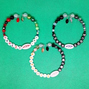 May include: Three beaded bracelets with white beads and colorful accents. The bracelets spell out the names "Harley Ivy", "Venom Eddie", and "Aziraphale Crowley".