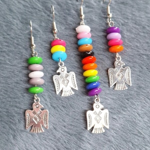 Könnte beinhalten: Drei Paar Hängeohrringe mit regenbogenfarbenen Perlen und einem silbernen Adler-Anhänger. Die Perlen sind in verschiedenen Farbkombinationen, darunter rosa, blau, grün und lila.