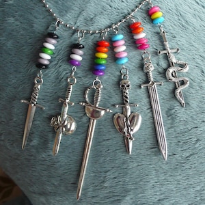 Puede incluir: Un collar de cadena de plata con cuentas de colores y dijes de espadas plateadas. Las cuentas están dispuestas en un patrón de arcoíris, con un patrón de cuentas blanco y negro a cada lado. Los dijes de espadas incluyen una daga, una espada con una calavera, una espada con un corazón, una espada con una serpiente y una espada con una calavera.