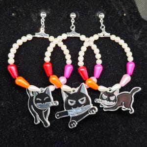 Puede incluir: Tres collares de perlas con dijes de gato negro. Cada collar tiene una cuenta de color diferente: naranja, rosa y rojo.