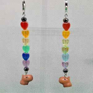 May include: Rainbow heart charm earrings with a capybara charm at the bottom. The earrings are made with silver-toned metal and have a rainbow of colors in the heart charms.