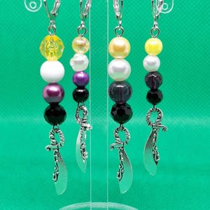 Puede incluir: Cuatro pares de pendientes colgantes con una variedad de cuentas de colores, incluyendo amarillo, blanco, morado y negro. Cada pendiente presenta un dije plateado en forma de espada curva. Los pendientes cuelgan de alambres plateados.