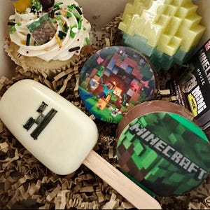 May include: A collection of Minecraft-themed treats including a cupcake with green and brown sprinkles, a white chocolate popsicle with a black H, a round chocolate with a game scene, a round chocolate with the Minecraft logo, and a block toy.