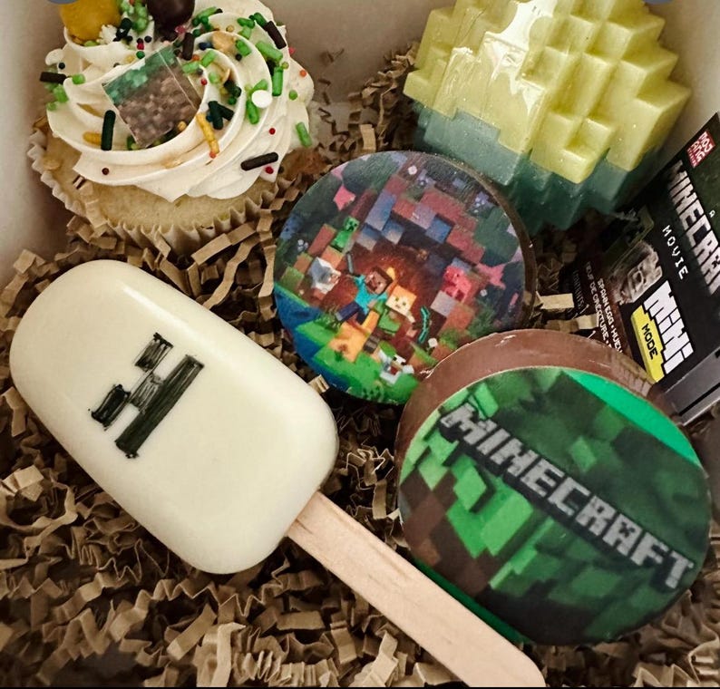 May include: A collection of Minecraft-themed treats including a cupcake with green and brown sprinkles, a white chocolate popsicle with a black H, a round chocolate with a game scene, a round chocolate with the Minecraft logo, and a block toy.