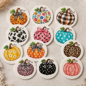 May include: A set of twelve round ceramic coasters, each featuring a unique pumpkin illustration. Designs include orange polka dots, floral patterns, black and white buffalo plaid, cow print, pink hearts, teal polka dots, leopard print, and leaf motifs.