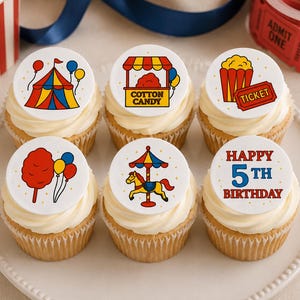 May include: Six cupcakes topped with white circular edible toppers featuring carnival-themed illustrations including a circus tent, cotton candy stand, popcorn, a ticket, a carousel horse, and text that reads Happy 5th Birthday in red and blue.