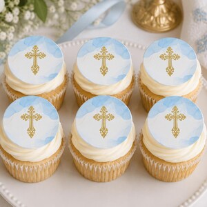 May include: Six cupcakes arranged on a white plate, each topped with a round edible topper featuring a gold cross design against a light blue watercolor background. The cupcakes have white frosting and are in white paper liners.