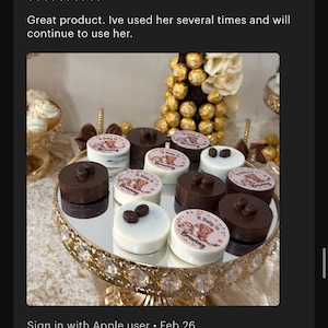May include: A collection of round chocolate treats arranged on a gold-rimmed mirrored pedestal stand. Some pieces are dark brown with coffee bean toppers, while others are white with a printed graphic featuring a teddy bear and the text A Baby Is Brewing.
