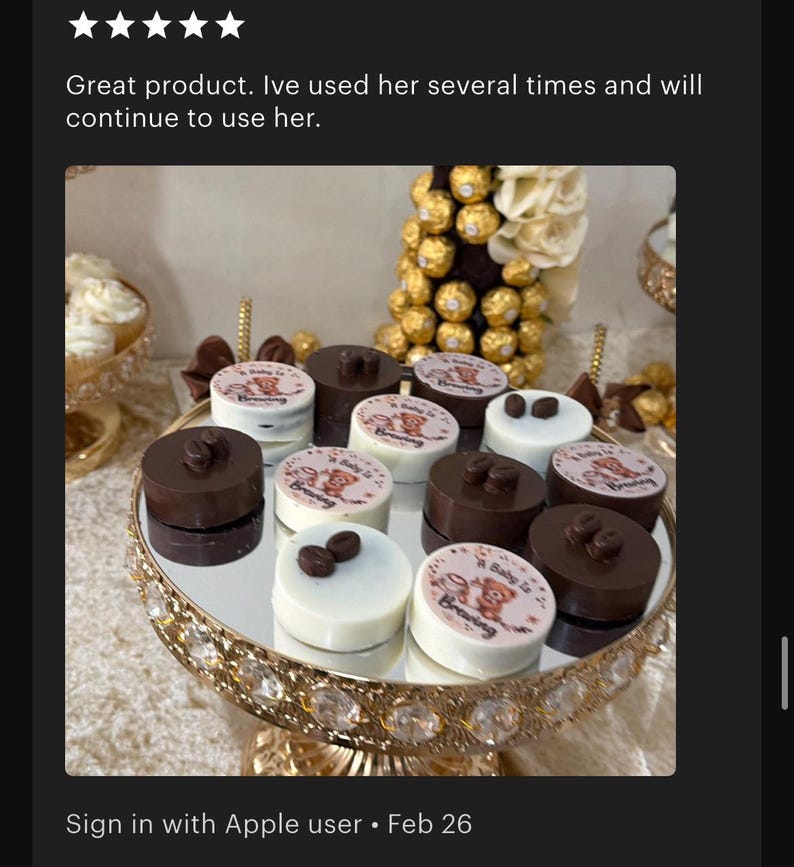 May include: A collection of round chocolate treats arranged on a gold-rimmed mirrored pedestal stand. Some pieces are dark brown with coffee bean toppers, while others are white with a printed graphic featuring a teddy bear and the text A Baby Is Brewing.