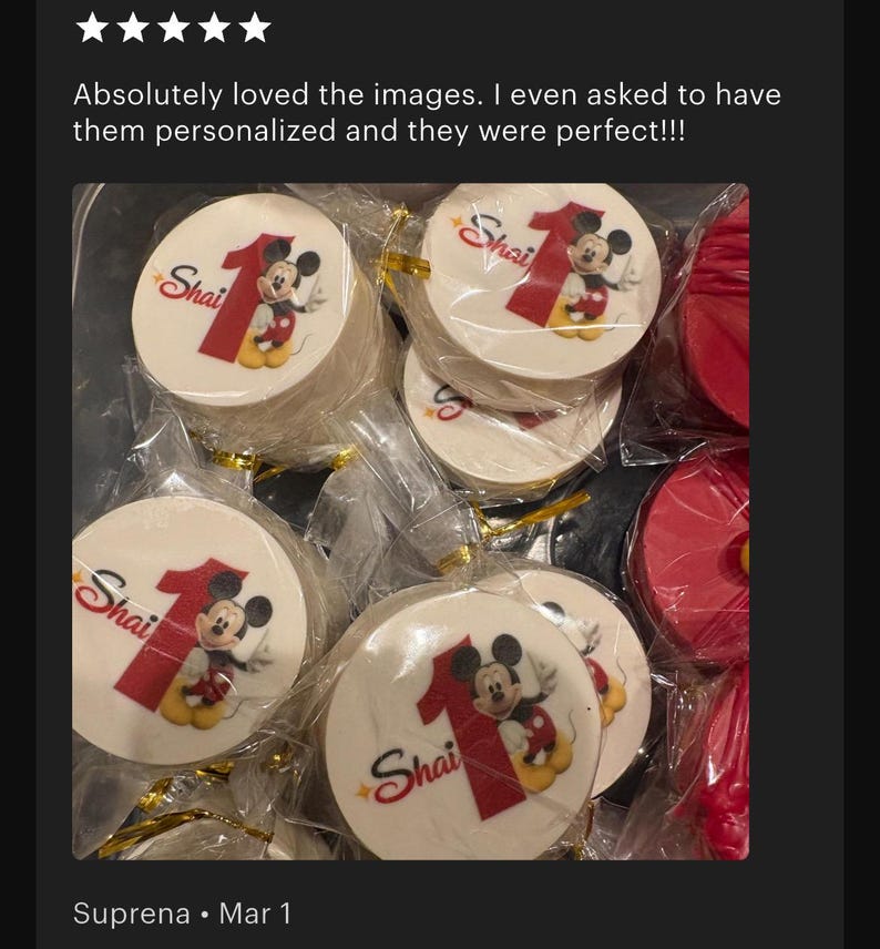 May include: Round white edible toppers individually wrapped in clear plastic, each featuring a red number one, the name Shai in red script, and a graphic of Mickey Mouse. The toppers are arranged in a group, with some red circular items visible.