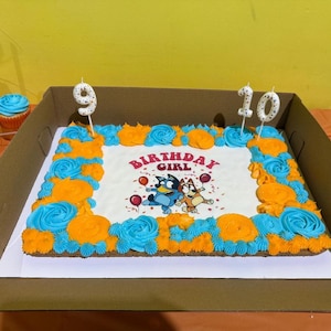 May include: A rectangular sheet cake with white frosting, decorated with a border of blue and orange piped rosettes. The center features an illustration of two cartoon dogs, balloons, and the text Birthday Girl. Two candles shaped as 9 and 10 are inserted.