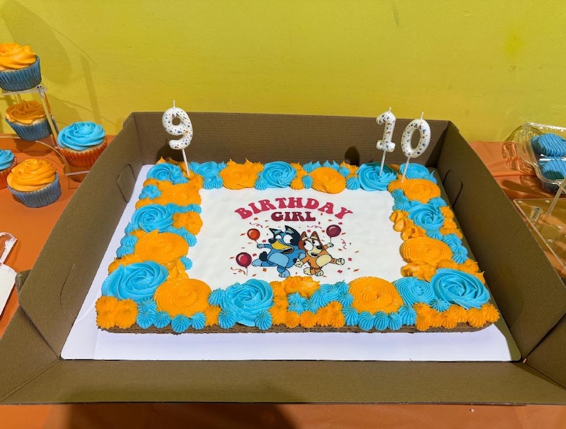 May include: A rectangular sheet cake with white frosting, decorated with a border of blue and orange piped rosettes. The center features an illustration of two cartoon dogs, balloons, and the text Birthday Girl. Two candles shaped as 9 and 10 are inserted.