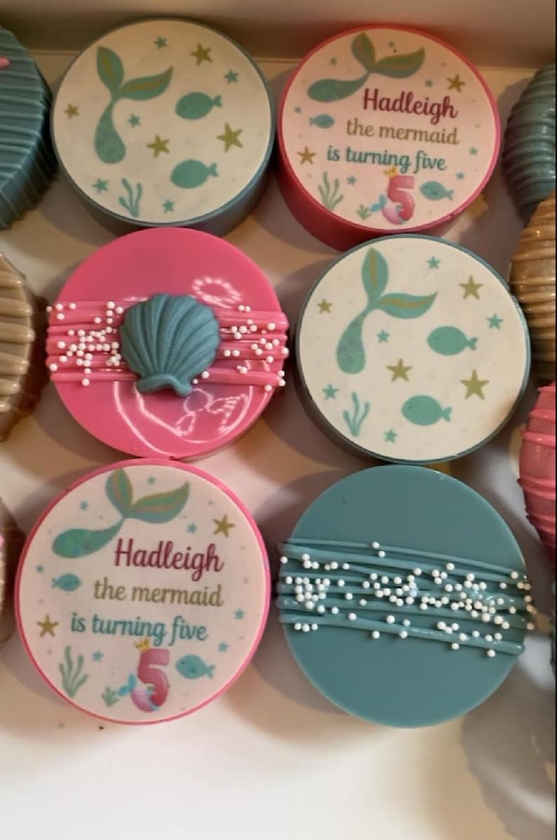 May include: A collection of round chocolate-covered cookies featuring mermaid-themed designs. Some cookies display the text Hadleigh the mermaid is turning five with fish and star graphics, while others feature a blue seashell or white sprinkles.