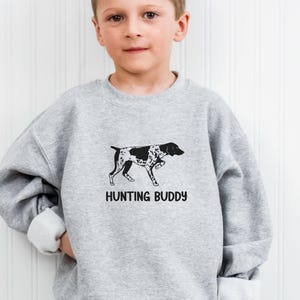 May include: A light gray crewneck sweatshirt featuring a black graphic of a spotted hunting dog in a standing position. Below the dog illustration, the text reads HUNTING BUDDY in a bold, black, sans-serif font. The garment has long sleeves.
