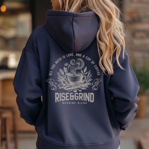 May include: Navy blue hoodie with a hood and a drawstring. The back features a coffee-themed graphic with the text "ALL YOU NEED IS LOVE, AND A CUP OF COFFEE" and "RISE&GRIND MORNING BLEND" in a light beige color. The design includes a coffee cup, leaves, and swirls.