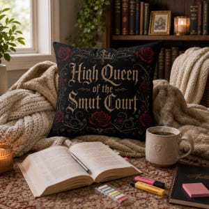 May include: Black throw pillow with the text "High Queen of the Smut Court" in a gothic font, surrounded by red roses and a crown. The pillow is on a cozy blanket with a book, coffee mug, and other items.