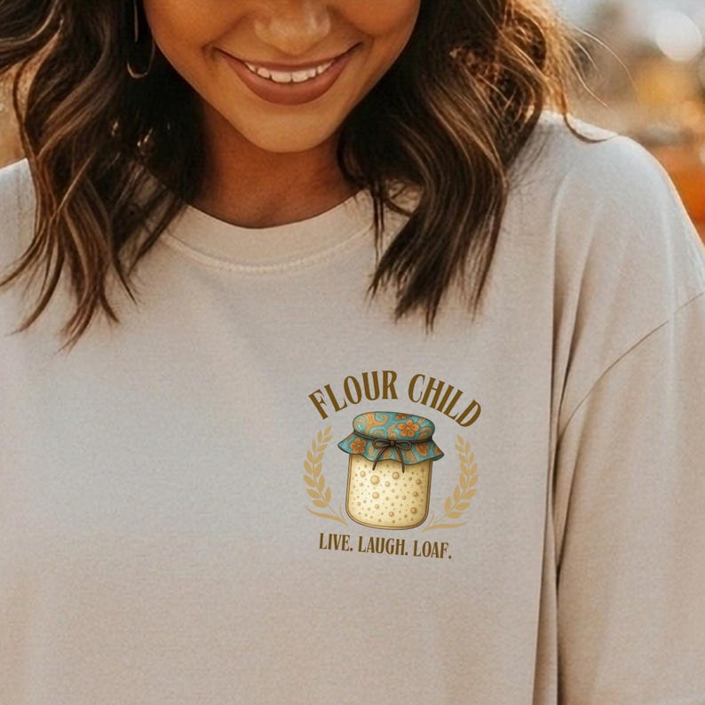 May include: A beige t-shirt featuring the text "FLOUR CHILD" and an illustration of a bread jar with a blue and orange lid. The jar is encircled by a wheat wreath, with the words "LIVE. LAUGH. LOAF." below the jar.