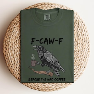 May include: Dark green t-shirt with the text "F-CAW-F" above a black raven illustration holding a coffee cup. The text "BEFORE I'VE HAD COFFEE" is below the raven. The shirt is folded on a woven surface.