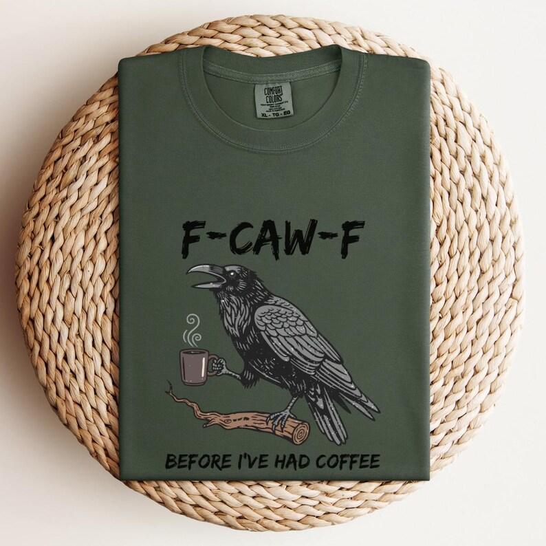 May include: Dark green t-shirt with the text "F-CAW-F" above a black raven illustration holding a coffee cup. The text "BEFORE I'VE HAD COFFEE" is below the raven. The shirt is folded on a woven surface.