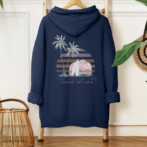 Op de afbeelding: Donkerblauwe hoodie met een grafische print van een vintage busje met surfplanken, palmbomen en een zonsondergang. De tekst "THE POGUE LIFE" is onder de print gedrukt, met daaronder de woorden "OUTER BANKS - NORTH CAROLINA".