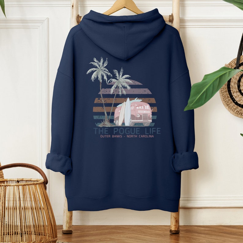 Op de afbeelding: Donkerblauwe hoodie met een grafische print van een vintage busje met surfplanken, palmbomen en een zonsondergang. De tekst "THE POGUE LIFE" is onder de print gedrukt, met daaronder de woorden "OUTER BANKS - NORTH CAROLINA".