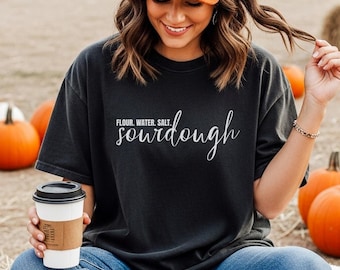 May include: Black t-shirt with the words "Flour, Water, Salt, sourdough" in white script. The person is wearing an orange cap and blue jeans. A coffee cup is held in the left hand. Pumpkins are in the background.