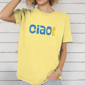 May include: A light yellow t-shirt with the word "Ciao" in blue and "Bella" in orange. The shirt has short sleeves and a relaxed fit. The model is wearing bracelets and blue jeans.